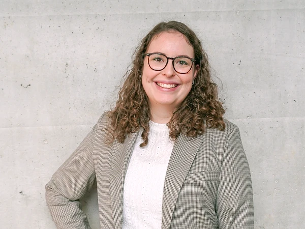 Eine lächelnde Frau mit lockigem Haar steht vor einer grauen Betonwand und trägt ein kariertes Blazer-Jackett über einem weißen Oberteil.