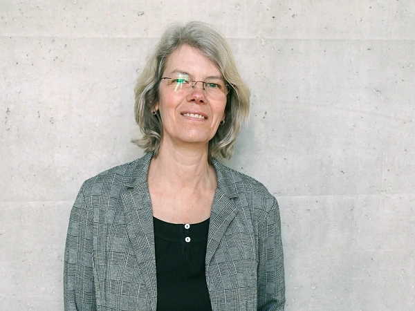 Eine lächelnde Frau mit blonden, schulterlangen Haaren trägt eine graue, kariierte Jacke und eine schwarze Bluse. Sie steht vor einer grauen Betonwand.