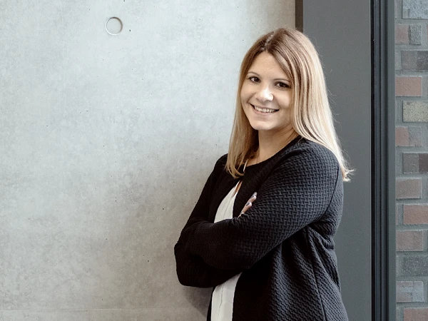 Eine lächelnde Frau steht mit verschränkten Armen vor einer minimalistischen, betonartigen Wand und schaut in die Kamera.