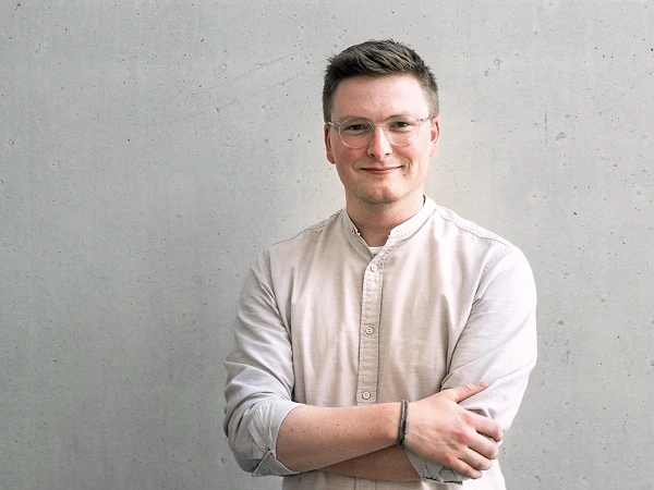 Ein junger Mann mit Brille trägt ein helles Hemd und steht mit verschränkten Armen vor einer grauen Betonwand.