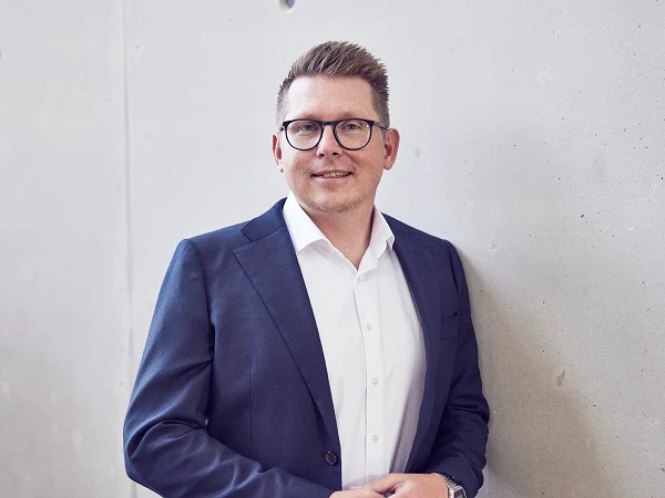 Ein Mann in einem Anzug mit einer weißen Bluse steht lächelnd an einer grauen Wand und hält seine Hände in einer entspannten Position.
