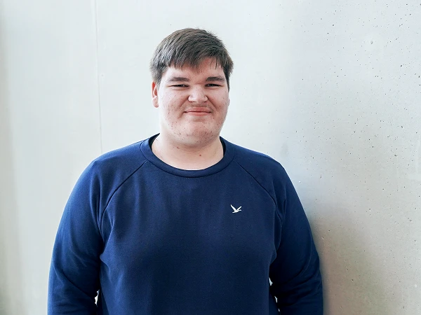 Ein junger Mann mit einem lächelnden Gesicht steht neben einer hellen, grauen Wand und trägt einen blauen Pullover mit einem kleinen Logo.