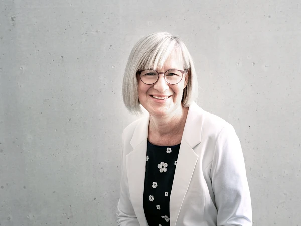 Eine lächelnde Frau mit kurzen, grauen Haaren und Brille trägt einen weißen Blazer über einem schwarzen, blumigen Oberteil und sitzt vor einer minimalistischen, grauen Wand.