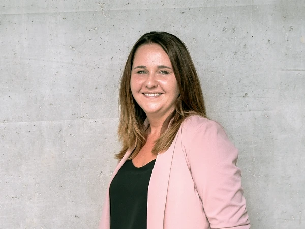 Eine lächelnde Frau mit langen, braunen Haaren steht vor einer grauen Betonwand. Sie trägt ein schwarzes Oberteil und einen rosafarbenen Blazer sowie eine hellblaue Jeans.