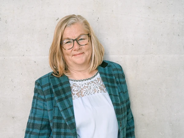 Eine lächelnde Frau mit mittellangen, blonden Haaren steht vor einer neutralen, hellgrauen Wand. Sie trägt eine karierten, blauen Blazer und eine helle Bluse mit Spitzenverzierungen.