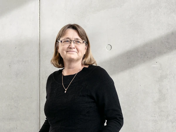 Eine lächelnde Frau mit schulterlangen, braunen Haaren trägt einen schwarzen Pullover und steht entspannt vor einer grauen Betonwand, während schräg ein Lichtstrahl auf die Wand fällt.