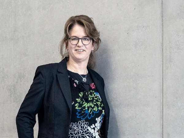 Eine Person steht lächelnd neben einer grauen Wand und trägt ein schwarzes Blazerjacke sowie ein blumiges Oberteil.