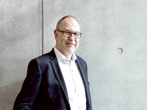 Ein Mann mit Brille und einem schwarzen Blazer steht lächelnd an einer grauen Betonnwand.