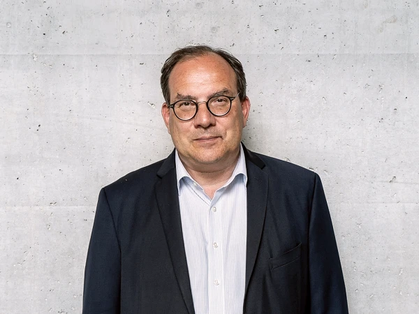 Ein Mann mit Brille steht vor einer grauen Betonnwand, trägt einen dunklen Anzug und ein helles Hemd und blickt direkt in die Kamera.