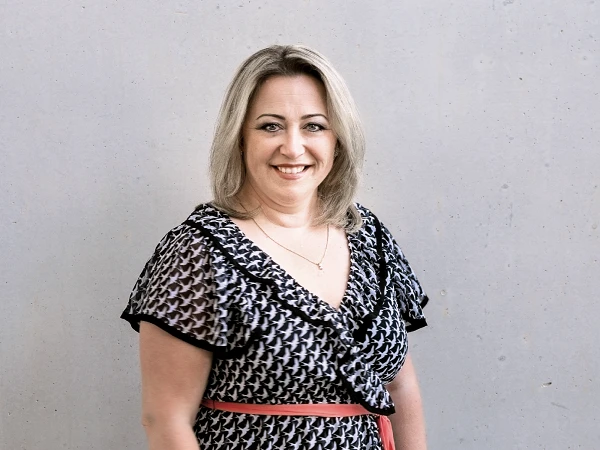 Eine lächelnde Frau mit blonden Haaren trägt ein gemustertes Kleid und steht vor einer grauen Wand.