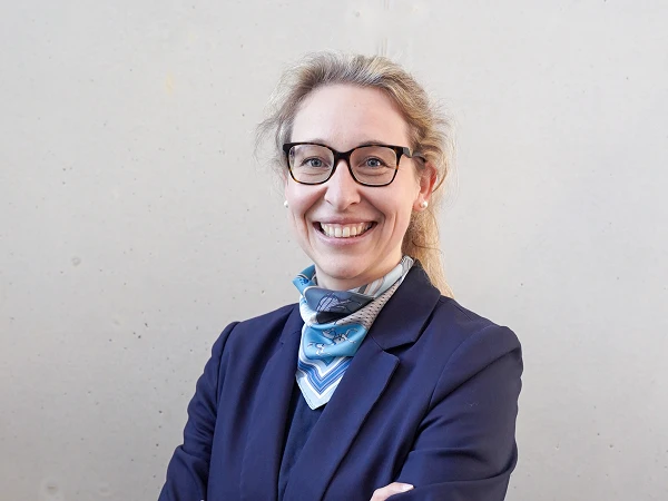 Eine lächelnde Frau mit Brille und Pferdeschwanz trägt einen blauen Blazer und einen bunten Schal. Sie steht mit verschränkten Armen vor einer grauen Wand.