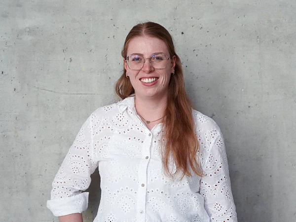 Eine lächelnde Frau mit langen roten Haaren trägt eine weiße Bluse mit Lochmuster und steht vor einer grauen Betonwand.