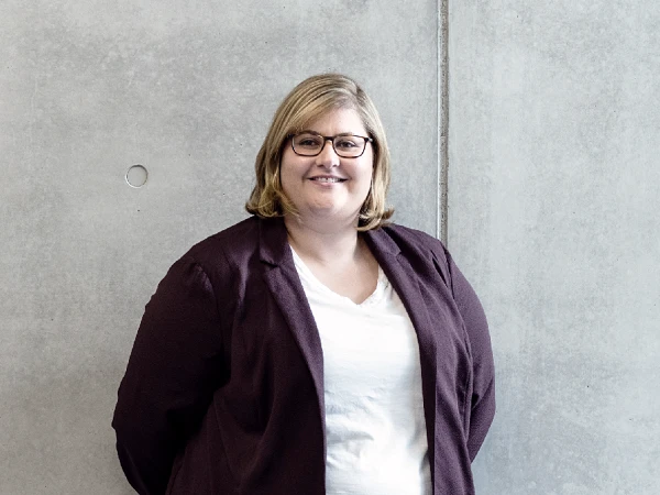 Eine lächelnde Frau mit blonden Haaren steht vor einer grauen, strukturierten Wand. Sie trägt eine dunkle Jacke über einem weißen T-Shirt und hat die Hände hinter dem Rücken verschränkt.