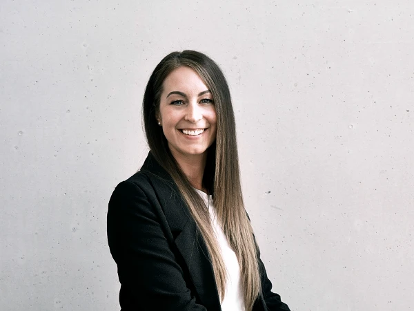 Eine lächelnde Frau mit langen, braunen Haaren sitzt auf einem Stuhl vor einer hellen, glatten Wand. Sie trägt einen schwarzen Blazer und ein weißes Oberteil und wirkt freundlich und selbstbewusst.