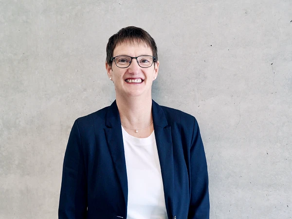 Eine lächelnde Frau mit kurzen, dunklen Haaren trägt ein schwarzes Blazer und ein weißes T-Shirt, steht vor einer grauen, strukturierten Wand.