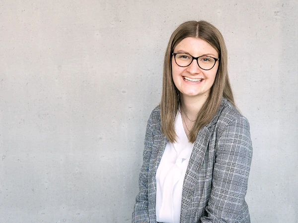 Eine junge Frau mit langen, braunen Haaren und einer Brille sitzt lächelnd vor einer grauen Betonwand, gekleidet in einem karierten Blazer und einem weißen Oberteil.