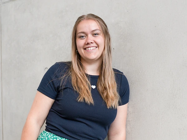 Eine junge Frau mit langen, hellen Haaren und einem lächelnden Gesicht steht lässig an einer grauen Wand, während sie ein schwarzes T-Shirt und einen grünen, blumigen Rock trägt.