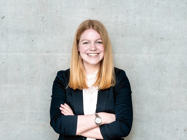 Eine junge Frau mit blonden Haaren steht mit verschränkten Armen vor einer grauen Betonwand und lächelt. Sie trägt einen schwarzen Blazer und ein weißes Oberteil.