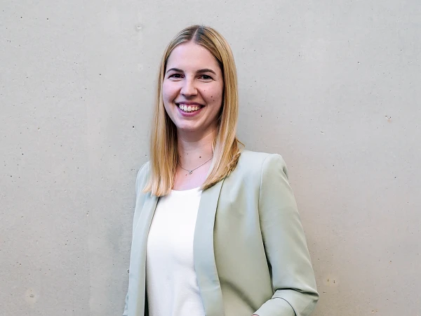 Eine lächelnde Frau mit langen, blonden Haaren trägt einen hellgrünen Blazer über einer weißen Bluse und steht vor einer grauen Wand.