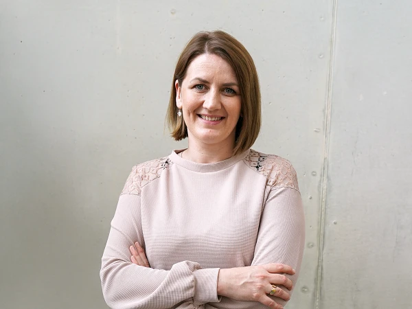 Eine lächelnde Frau mit kurzem, braunem Haar steht mit verschränkten Armen vor einer grauen Wand. Sie trägt ein helles Oberteil mit transparenten Ärmeln und wirkt freundlich und einladend.