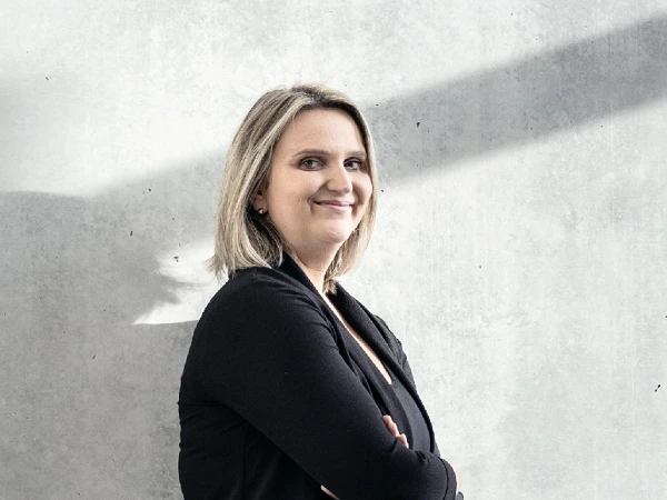 Eine lächelnde Frau steht mit verschränkten Armen vor einer grauen Betonwand, die von sanftem Licht durch einen Fensterstrahl beleuchtet wird.