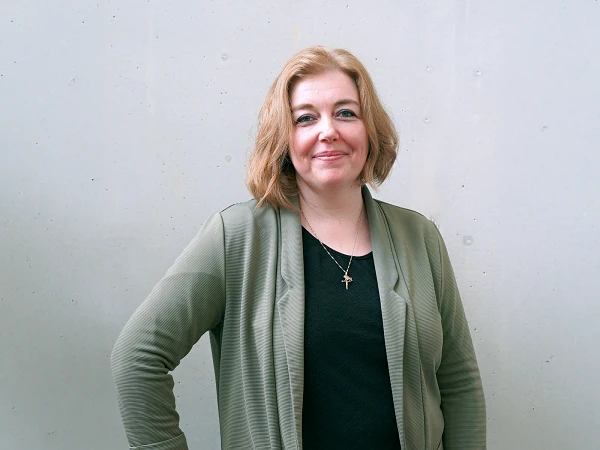 Eine Dame mit schulterlangen, blonden Haaren steht vor einer grauen Wand. Sie trägt eine olivegrüne Jacke und ein schwarzes Oberteil, hat die rechte Hand in die Hüfte gestützt und lächelt freundlich in die Kamera.