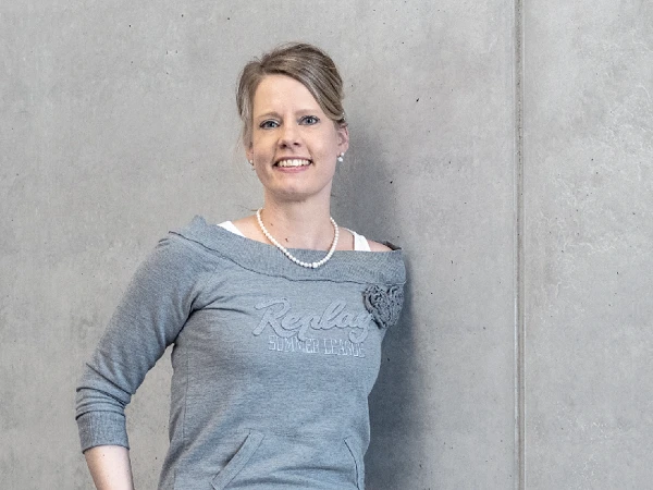 Eine Frau in einem grauen Sweatshirt mit Schriftzug steht lächelnd gegen eine graue Wand. Sie trägt eine Perlenkette und hat ihre Haare locker frisiert.