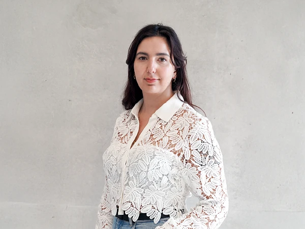 Eine Frau mit langen, dunklen Haaren trägt eine transparente, weiße Spitzenbluse und steht vor einem grauen Hintergrund. Sie blickt direkt in die Kamera.