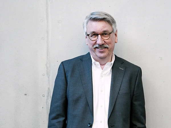 Ein lächelnder Mann mit Brille und einem grauen Anzug steht vor einer hellen, glatten Wand.