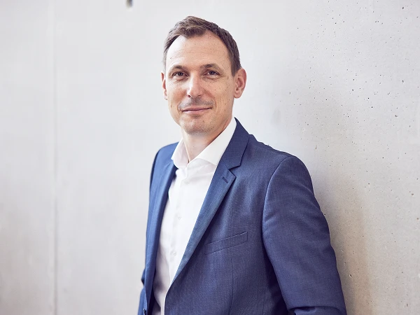 Ein mittelgroßer Mann in einem blauen Anzug mit weißem Hemd lächelt entspannt, während er an einer grauen Wand steht.