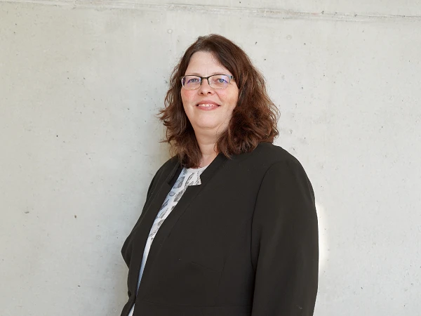 Eine Frau mit lockigem braunem Haar und Brille lächelt vor einer neutralen Betonwand, gekleidet in eine schwarze Jacke.