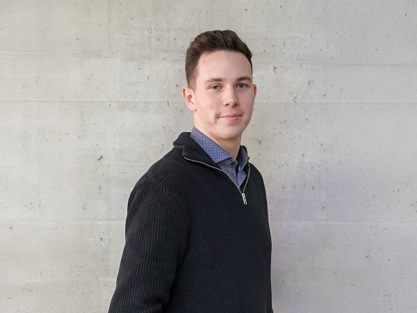 Ein junger Mann steht seitlich vor einer grauen Betonwand, trägt einen schwarzen Pullover mit Reißverschluss und ein kariertes Hemd.