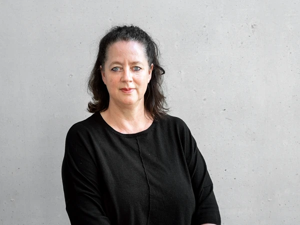 Eine Frau mit dunklen, schulterlangen Haaren trägt ein schwarzes Oberteil und steht vor einer grauen, strukturierten Wand. Sie schaut direkt in die Kamera und hat einen neutralen Gesichtsausdruck.