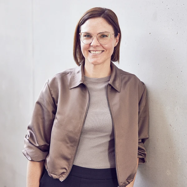 Eine lächelnde Frau mit Brille steht entspannt mit einer Hand in der Hosentasche an einer hellgrauen Wand. Sie trägt eine braune Jacke über einem hellen Oberteil und hat schulterlange, brünette Haare.