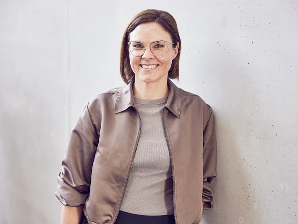 Eine lächelnde Frau mit Brille steht entspannt mit einer Hand in der Hosentasche an einer hellgrauen Wand. Sie trägt eine braune Jacke über einem hellen Oberteil und hat schulterlange, brünette Haare.