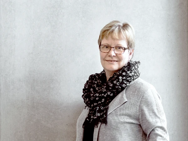 Eine lächelnde Frau mit kurzen blonden Haaren und einer Brille steht vor einer grauen Wand, während sie einen schwarzen Schal mit Muster trägt.