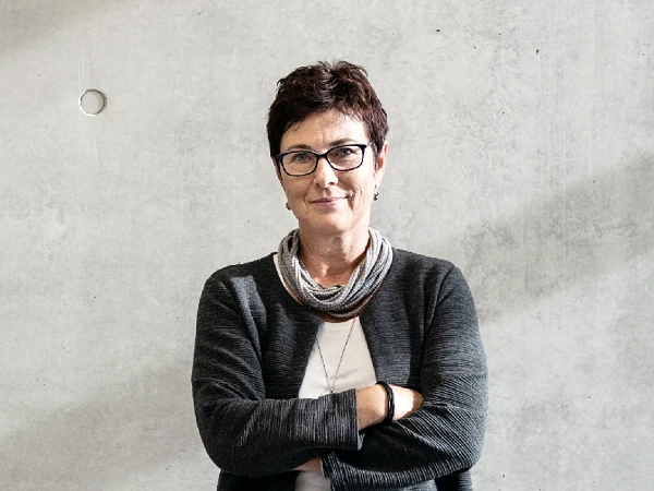 Eine lächelnde Frau mit kurzen, braunen Haaren und Brille steht mit verschränkten Armen vor einer grauen Wand. Sie trägt ein schwarzes Oberteil und eine graue Strickjacke, dazu einen hellen Schal.