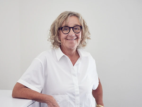 Eine lächelnde ältere Frau mit lockigem Haar steht an einer Wand und lehnt sich leicht an einen weißen Schrank. Sie trägt eine weiße Bluse und hat eine Brille auf.