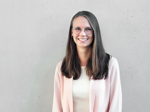Eine lächelnde Frau mit langen, glatten Haaren und Brille steht vor einer grauen Betonwand. Sie trägt ein helles Oberteil und einen blassen Blazer.