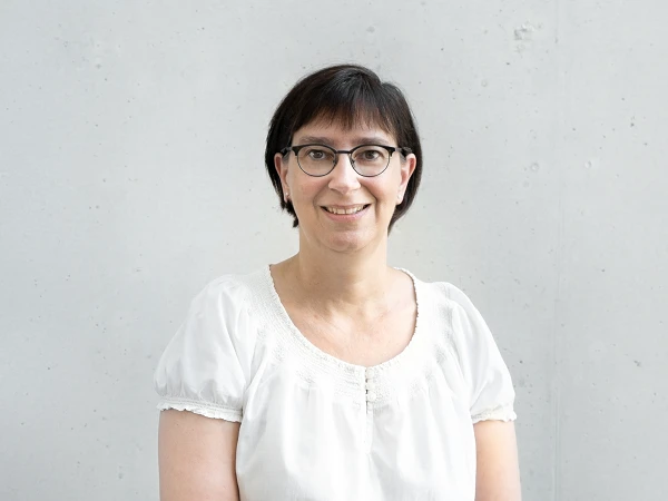 Eine Frau mit kurzer, dunkler Frisur und Brille lächelt freundlich vor einer grauen Betonwand, während sie ein weißes Oberteil trägt.