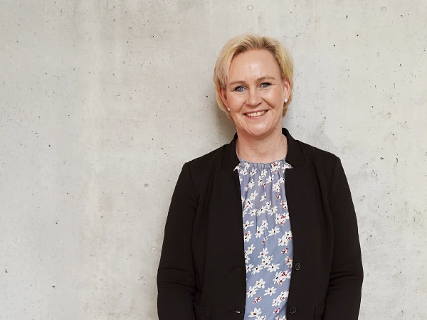 Eine lächelnde Frau mit blonden Haaren trägt eine schwarze Blazerjacke über einem blauen, blümchenmusterigen Oberteil und steht vor einer grauen, strukturierten Wand.