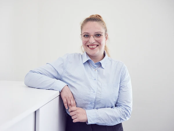 Eine lächelnde Frau mit blonden Haaren, die zu einem Pferdeschwanz gebunden sind, trägt eine hellblaue Bluse und steht entspannt an einem weißen Regal in einem hellen Raum.
