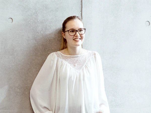 Eine lächelnde Frau mit Brille steht an einer grauen Wand, trägt eine weiße Bluse mit feinen Details und hat ihre Hände in die Hüften gestützt.