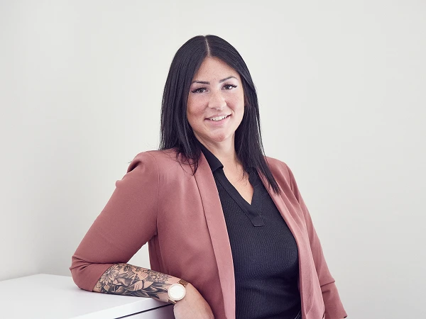 Eine Frau mit langen, schwarzen Haaren und einem eleganten, rosafarbenen Blazer steht an einer weißen Kommode und lächelt in die Kamera. Sie trägt ein schwarzes oberteil und hat mehrere Tattoos an ihrem linken Arm.