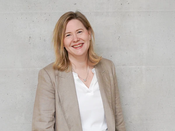 Eine blonde Frau in einem beigen Blazer und weißem Oberteil lächelt vor einer grauen Betonwand.