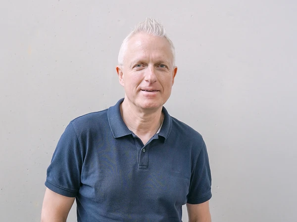 Ein Mann mit grauen, kurzen Haaren steht vor einer hellgrauen Wand und lächelt, während er ein dunkelblaues Poloshirt trägt.