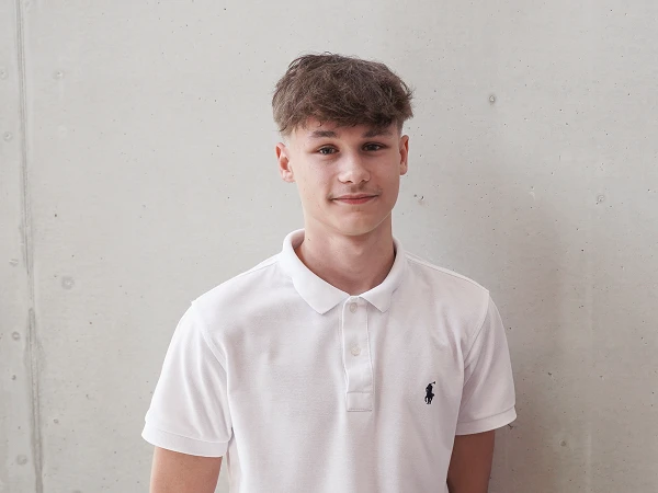 Ein junger Mann mit kurzem, braunem Haar trägt ein weißes Polo-Shirt und steht vor einer glatten, hellgrauen Wand.