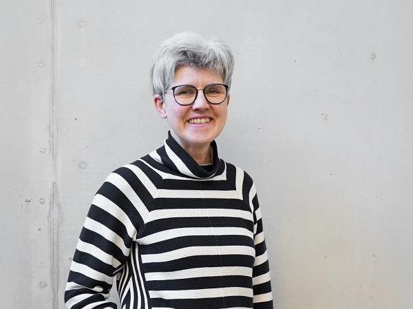Eine lächelnde Frau mit kurzen grauen Haaren trägt einen schwarz-weiß gestreiften Pullover und steht vor einer neutralen, hellgrauen Wand.