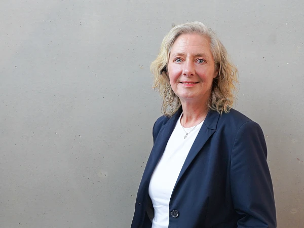 Eine lächelnde Frau mit lockigem, blondem Haar steht vor einer grauen Wand und trägt einen dunkelblauen Blazer über einem weißen Oberteil.