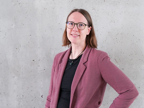 Eine Person mit braunen Haaren und Brille steht vor einer grauen Betonwand und trägt einen lila Blazer über einem schwarzen Oberteil. Sie lächelt leicht und hat eine Hand an die Hüfte gelegt.
